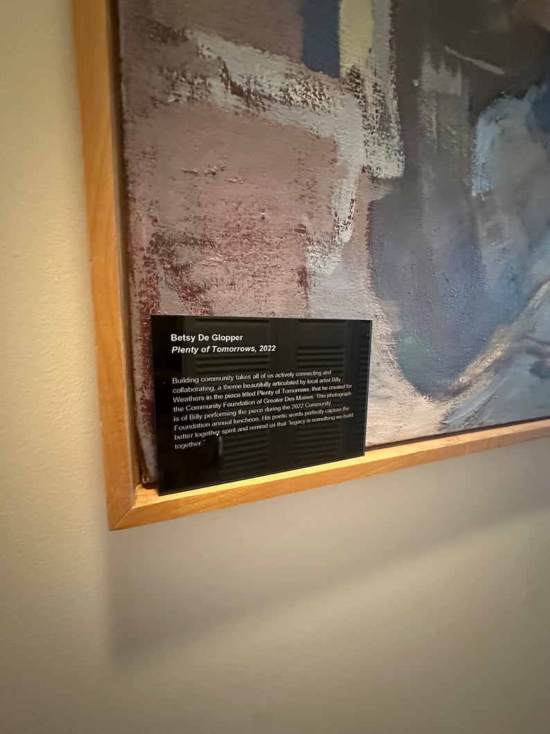 May include: A black plaque with white text on a wooden frame. The text reads "Betsy De Glopper, Plenty of Tomorrows, 2022". The plaque is mounted on a wall next to an abstract painting with shades of gray, pink, and white.