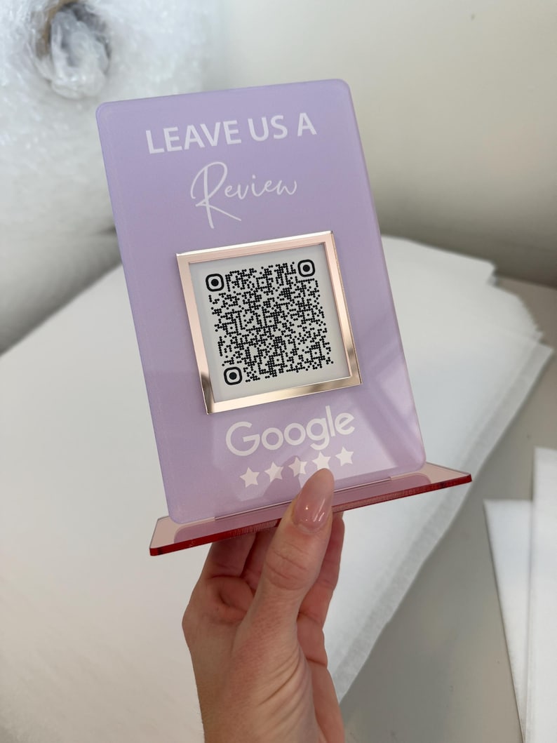K&ouml;nnte beinhalten: Ein lilafarbenes Acrylschild mit dem Text "LEAVE US A Review" und einem goldgerahmten QR-Code. Das Schild zeigt auch das Google-Logo und Sternebewertungen. Der Sockel des Schildes ist transparent rot.