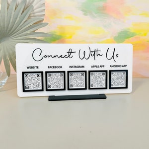 May include: White sign with the text "Connect With Us" and five black squares containing QR codes labeled "Website", "Facebook", "Instagram", "Apple App", and "Android App".