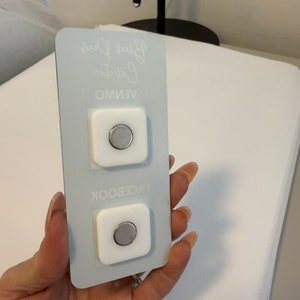 May include: Two white square magnetic phone mounts with a silver magnetic disc in the center. The mounts are on a light blue background with the text "VENMO", "Collective", and "Black Pearl" written in white.