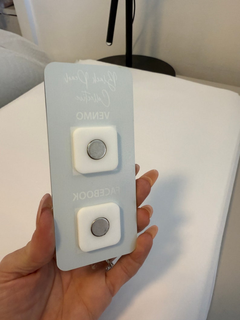 May include: Two white square magnetic phone mounts with a silver magnetic disc in the center. The mounts are on a light blue background with the text "VENMO", "Collective", and "Black Pearl" written in white.