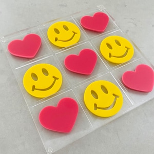 May include: A clear plastic game board with nine spaces. Each space contains either a yellow smiley face or a pink heart. The board is on a gray surface.