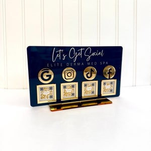 May include: A black sign with gold lettering that reads "Let's Get Social" and "Elite Derma Med Spa". The sign has four gold icons for social media platforms: Google, Instagram, TikTok, and Facebook. Each icon has a QR code underneath it.