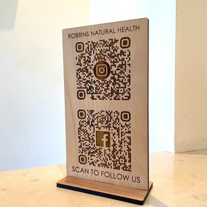 May include: Wooden sign with the text "ROBBINS NATURAL HEALTH" and two QR codes. The top QR code has an Instagram logo and the bottom QR code has a Facebook logo. The text "SCAN TO FOLLOW US" is below the QR codes.