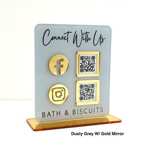 May include: A dusty grey acrylic sign with a gold mirror base. The sign says "Connect With Us" and features icons for Facebook, Instagram, and QR codes. The text "Bath & Biscuits" is also on the sign.