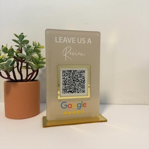 K&ouml;nnte beinhalten: Ein klarer Acrylst&auml;nder mit dem Schriftzug &bdquo;LEAVE US A Review&ldquo;. Ein QR-Code ist unter dem Text zentriert, mit dem Google-Logo und f&uuml;nf goldenen Sternen unten. Links ist eine kleine Topfpflanze.