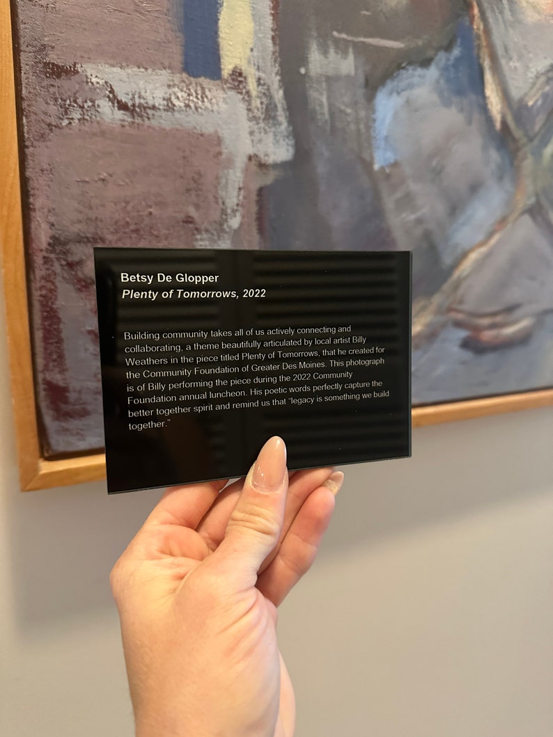 May include: A black plaque with white text on a wooden frame. The text reads "Betsy De Glopper, Plenty of Tomorrows, 2022." The text continues, "Building community takes all of us actively connecting and collaborating, a theme beautifully articulated by local artist Billy Weathers in the piece titled Plenty of Tomorrows, that he created for the Community Foundation of Greater Des Moines. This photograph is of Billy performing the piece during the 2022 Community Foundation annual luncheon. His poetic words perfectly capture the better together spirit and remind us that legacy is something we build together."