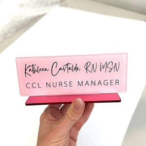 May include: Pink acrylic desk nameplate with the text "Kathleen Castaldo, RN MSN CCL Nurse Manager" in black lettering.