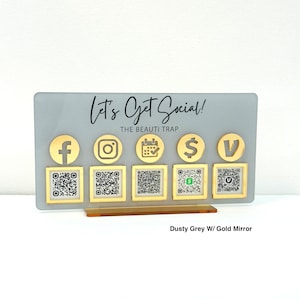 May include: A gray acrylic sign with a gold mirror base. The sign says "Let's Get Social! The Beauti Trap" and has icons for Facebook, Instagram, a calendar, a dollar sign, and a check mark. Below the icons are QR codes for each social media platform.