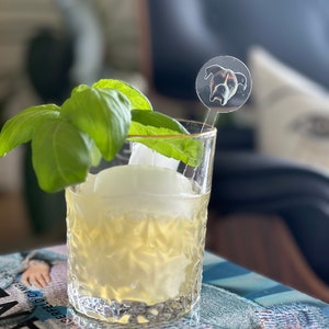 May include: A clear glass with a yellow liquid and ice inside. A sprig of green basil is on the rim of the glass. A clear plastic drink stirrer with a round, clear plastic topper with a printed image of a dog's face is in the glass.