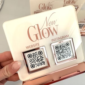 May include: A white acrylic sign with the text "New Glow" in rose gold lettering. The sign has two QR codes, one labeled "Website" and the other labeled "Instagram".