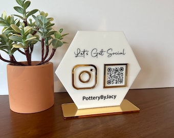 May include: White hexagonal sign with gold trim, featuring a black Instagram logo and a black and white QR code. The sign reads "Let's Get Social" and "PotteryByJacy".