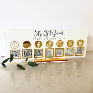 May include: A white rectangular acrylic sign with the text Let's Get Social and ANGIE'S DANCE PLUS. It features seven gold circular social media icons above seven square QR codes, including symbols for a website, Remind, Facebook, TikTok, Instagram, Cash App, and Venmo.
