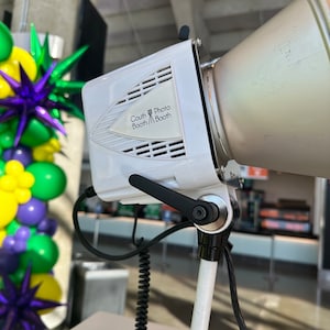 May include: White photo booth light with a white reflector and a black handle. The light has a label that says "South Photo Booth Booth".