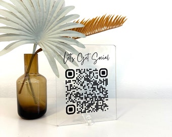 May include: A clear acrylic sign with the text "Let's Get Social" and a black and white QR code. The sign is on a white surface.