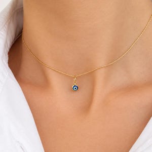 May include: A delicate gold chain necklace featuring a small, circular evil eye pendant. The pendant consists of a gold rim surrounding a blue, white, and black eye motif. The necklace rests against the neck, showcasing a simple, refined design.