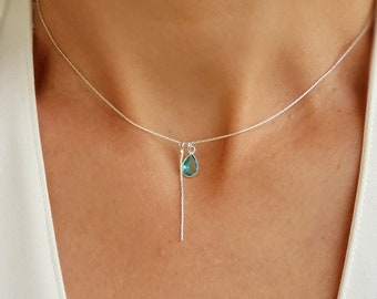 May include: A delicate silver necklace featuring a teardrop-shaped, teal-colored gemstone pendant. A vertical silver bar hangs below the pendant. The necklace is worn against a white top, showcasing the jewelry's minimalist design.