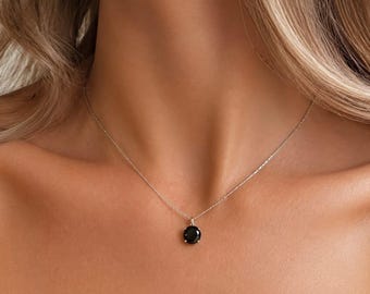 May include: A delicate silver-toned chain necklace featuring a single, round, faceted black gemstone pendant held in a four-prong setting. The necklace rests against the collarbone, showcasing a minimalist design with a polished metallic finish.