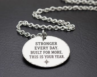 May include: A silver-toned circular pendant necklace featuring a hammered edge and an engraved message that reads: STRONGER EVERY DAY. BUILT FOR MORE. THIS IS YOUR YEAR. A small four-pointed star symbol is centered below the text on the metal disc.