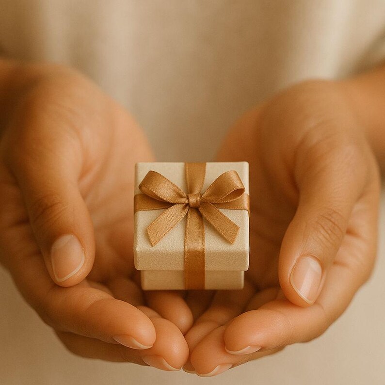 May include: A small, square, beige gift box featuring a smooth, satin-finish ribbon tied in a neat bow across the top. The box has a clean, minimalist design with a matte texture, presenting a neutral color palette that highlights the ribbon.