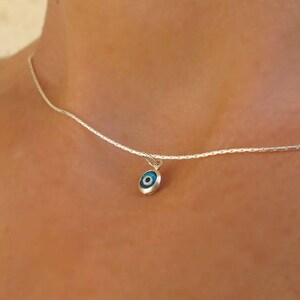 May include: A delicate silver chain necklace featuring a small, circular pendant. The pendant is a blue, white, and black evil eye charm with a metallic silver border, suspended from a simple jump ring attached to the thin, textured silver chain.