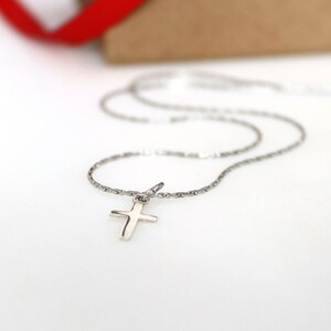 May include: A silver-toned metal necklace featuring a small, plain cross pendant suspended from a delicate, textured chain. The cross has a smooth, polished finish and is attached to the chain by a simple circular jump ring connector.