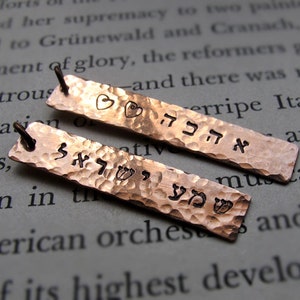 May include: Two hammered copper pendants with Hebrew text and heart and flower designs. The pendants have a textured surface and small loops for hanging. The background is a page of printed text.