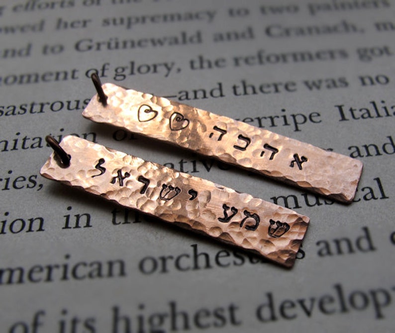 May include: Two hammered copper pendants with Hebrew text and heart and flower designs. The pendants have a textured surface and small loops for hanging. The background is a page of printed text.