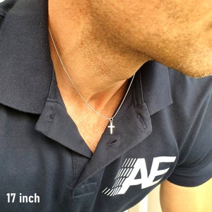 May include: A silver-toned metal cross pendant suspended on a thin, textured chain. The necklace is labeled as 17 inches in length. The cross features a simple, clean design with a polished finish, hanging centered against a dark blue polo shirt.