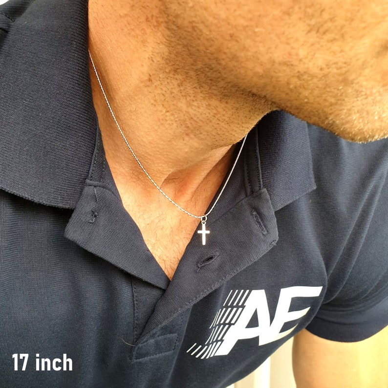 May include: A silver-toned metal cross pendant suspended on a thin, textured chain. The necklace is labeled as 17 inches in length. The cross features a simple, clean design with a polished finish, hanging centered against a dark blue polo shirt.