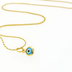 May include: A gold-toned chain necklace featuring a small, circular pendant with an evil eye motif. The pendant displays a central black pupil surrounded by a light blue iris and a white ring, all encased within a polished gold-colored metal frame.