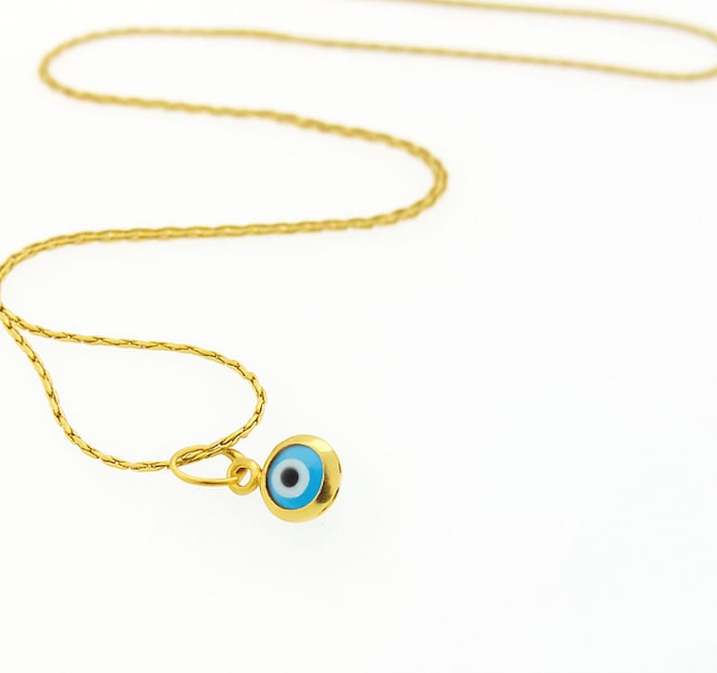 May include: A gold-toned chain necklace featuring a small, circular pendant with an evil eye motif. The pendant displays a central black pupil surrounded by a light blue iris and a white ring, all encased within a polished gold-colored metal frame.