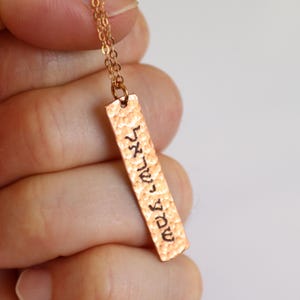 May include: A gold-colored necklace with a rectangular pendant. The pendant has a hammered texture and features Hebrew text. The chain is delicate, and the pendant hangs vertically. The necklace is displayed against a neutral background.
