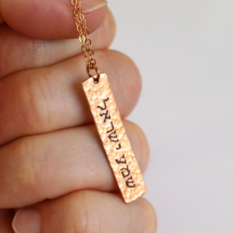 May include: A gold-colored necklace with a rectangular pendant. The pendant has a hammered texture and features Hebrew text. The chain is delicate, and the pendant hangs vertically. The necklace is displayed against a neutral background.