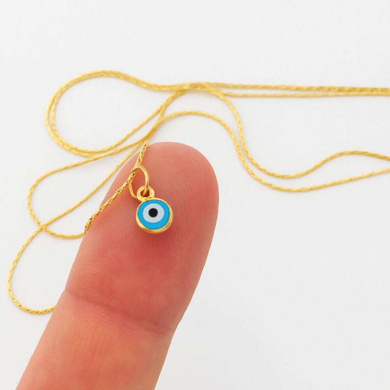 May include: A gold chain necklace featuring a small, circular pendant with an evil eye design. The pendant has a gold rim, a light blue outer ring, a white middle ring, and a black center dot. The delicate gold chain is thin and metallic.