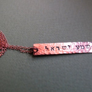 May include: A copper-colored necklace with a rectangular pendant. The pendant has hammered texture and features Hebrew text. The chain is also copper-colored. The necklace is displayed against a dark green background.