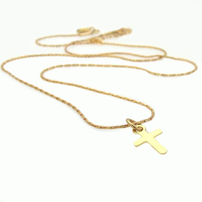 May include: A gold-toned metal necklace featuring a simple, plain cross pendant suspended from a delicate, textured chain. The cross has smooth, flat surfaces and rounded edges, while the chain consists of small, interconnected links.