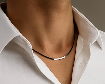 May include: A black cord necklace featuring a single, smooth, polished silver-toned metal bar pendant centered on the cord. The necklace is worn around the neck, resting against the collar of a crisp, white, button-down collared shirt.