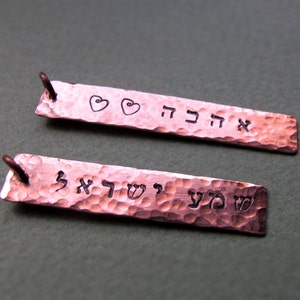 May include: Two copper rectangular pendants with hammered textures. One pendant has the word "אהבה" and two heart symbols. The other pendant has the words "שמע ישראל". Both pendants have a small loop at the top.