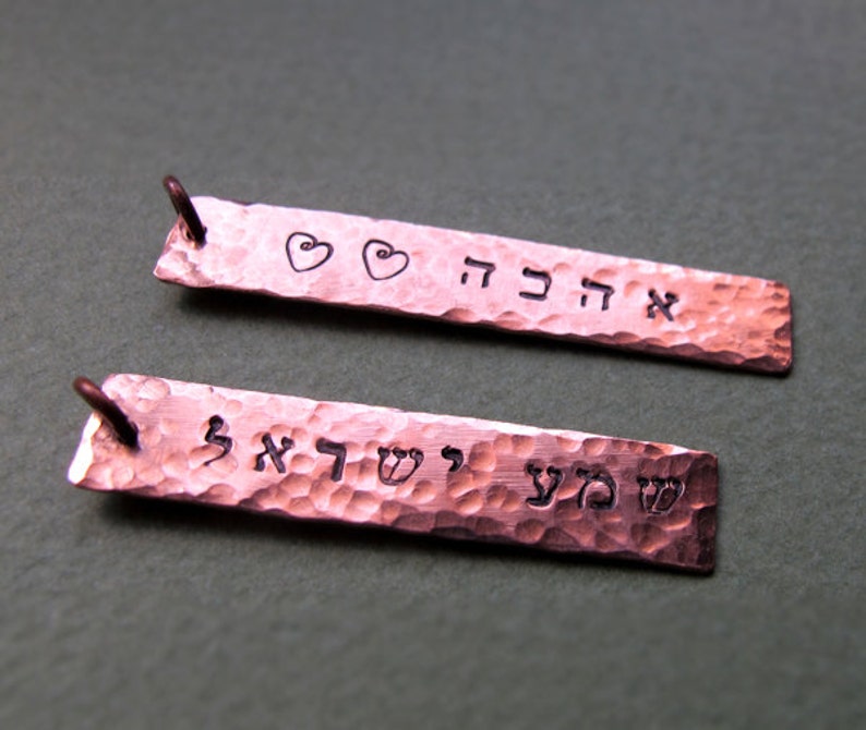 May include: Two copper rectangular pendants with hammered textures. One pendant has the word "אהבה" and two heart symbols. The other pendant has the words "שמע ישראל". Both pendants have a small loop at the top.