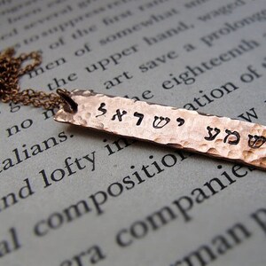 May include: A copper-colored, hammered metal pendant necklace. The rectangular pendant features stamped Hebrew characters and hangs from a delicate chain. The pendant's surface has a textured, dimpled appearance.