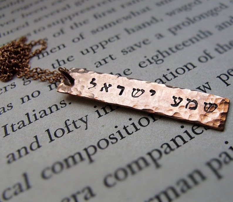 May include: A copper-colored, hammered metal pendant necklace. The rectangular pendant features stamped Hebrew characters and hangs from a delicate chain. The pendant's surface has a textured, dimpled appearance.