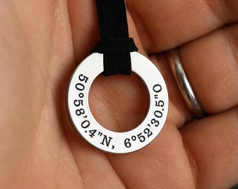 May include: A circular silver pendant featuring the engraved coordinates 50°58'0.4"N, 6°52'30.5"O, suspended from a black cord. The metallic ring has a polished finish and displays the geographic location clearly etched along its front surface.