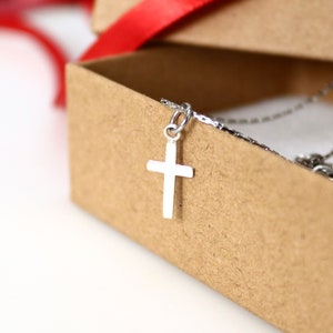 May include: A silver-toned cross pendant hangs from a delicate chain, resting against the edge of a small, light brown cardboard box. The cross features a simple, smooth design with a polished finish, suspended by a small circular jump ring.