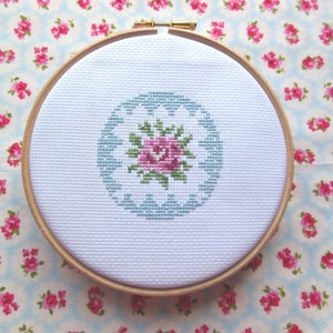 May include: A white embroidery hoop with a light blue and pink floral design. The design features a single pink rose with green leaves and a light blue circular border.