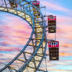 May include: A large Ferris wheel with red passenger cars against a colorful sunset sky. The wheel is made of metal and has a white background. The cars are suspended from the wheel by cables.