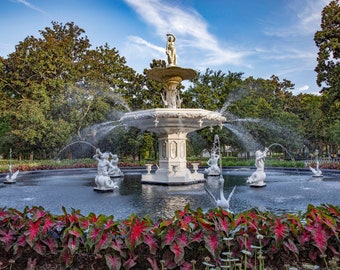 May include: A white fountain with multiple tiers and sculptures of people and birds. The fountain is surrounded by lush green trees and red foliage.