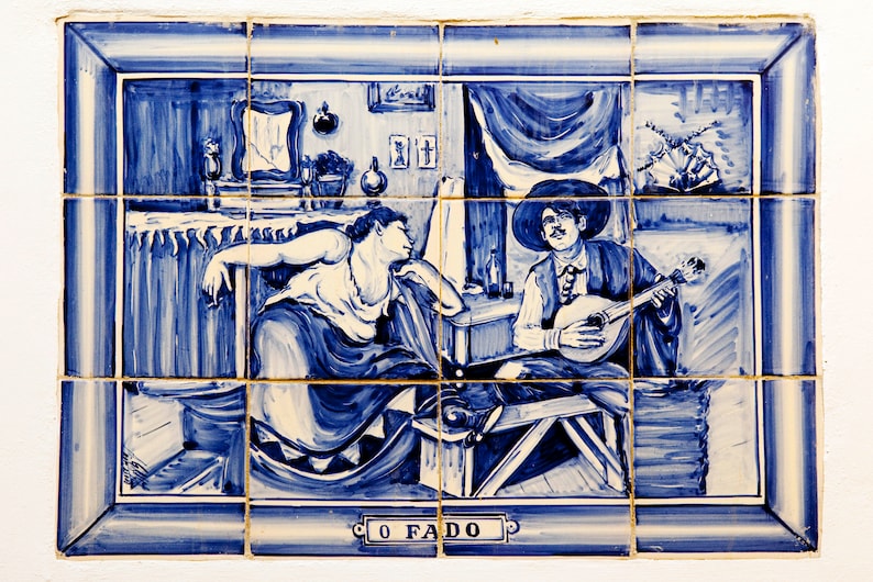 May include: A blue and white tile painting depicting a woman and a man playing a stringed instrument. The woman is wearing a long dress and the man is wearing a hat and a long coat. The painting is titled "O Fado".