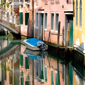 May include: A small blue and white boat is docked in a canal in Venice, Italy. The boat is covered with a blue tarp and has a small motor. The canal is lined with colorful buildings with windows and doors. The water is calm and reflects the buildings and sky.