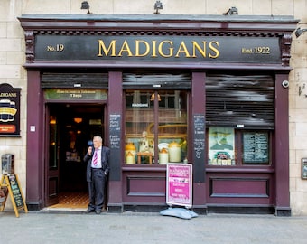 May include: A brown brick building with a dark brown awning that reads "Madigans" in gold letters. The awning has the number "19" and the text "Estd. 1922". A man in a suit stands in the doorway of the building.